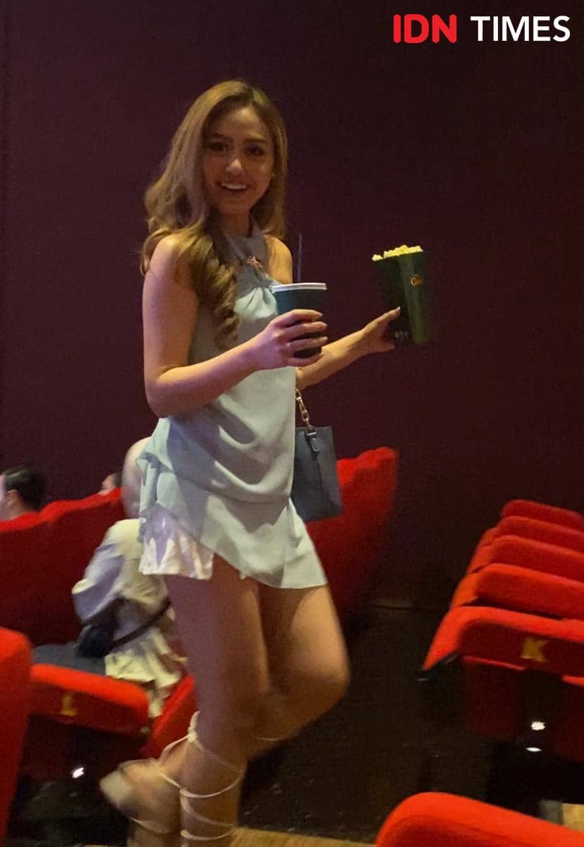 Seorang wanita berbusana biru muda membawa popcorn dan minuman di dalam bioskop dengan kursi merah saat acara special screening Moana live-action di Jakarta.