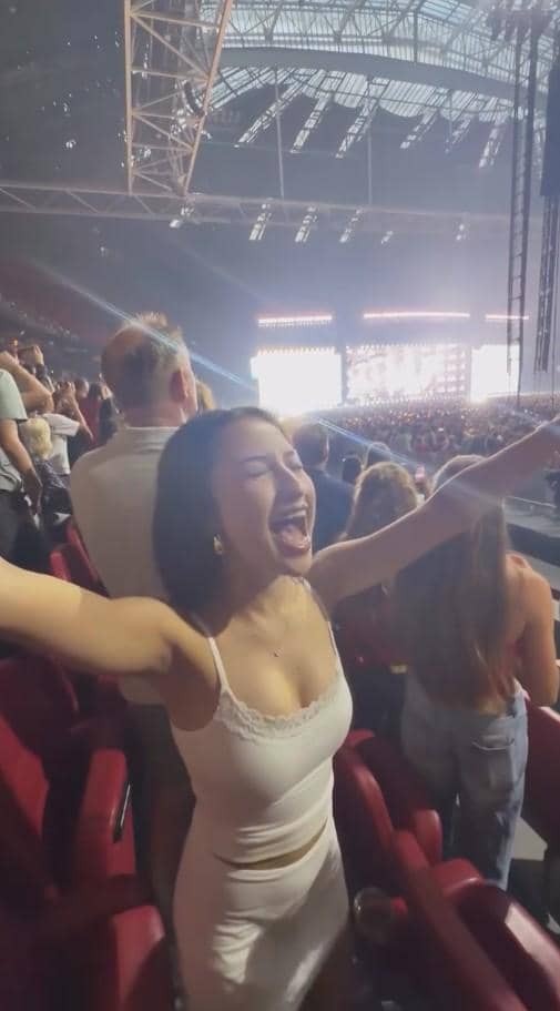 Seorang perempuan mengenakan gaun putih tersenyum lebar dan mengangkat tangan saat menikmati konser di stadion besar yang penuh penonton.