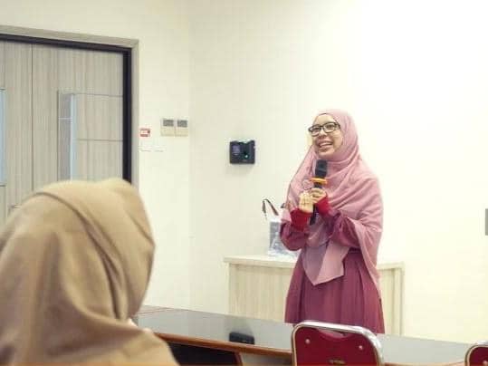 Seorang perempuan berhijab berbicara sambil memegang mikrofon di depan peserta dalam ruangan kelas dengan suasana akademik.