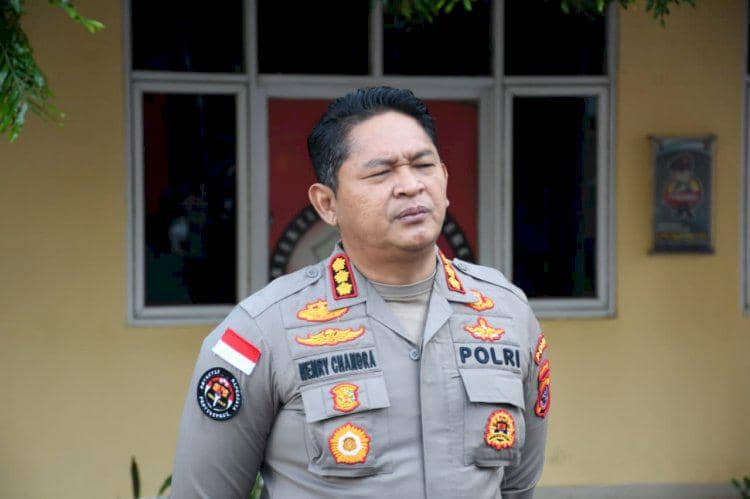 Seorang perwira polisi mengenakan seragam lengkap dengan berbagai lencana dan atribut kepolisian berdiri di depan gedung berlogo Polri.