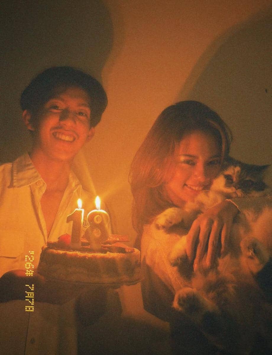 Seorang perempuan memegang kucing sambil tersenyum di samping seorang laki-laki yang membawa kue ulang tahun dengan lilin angka 18.