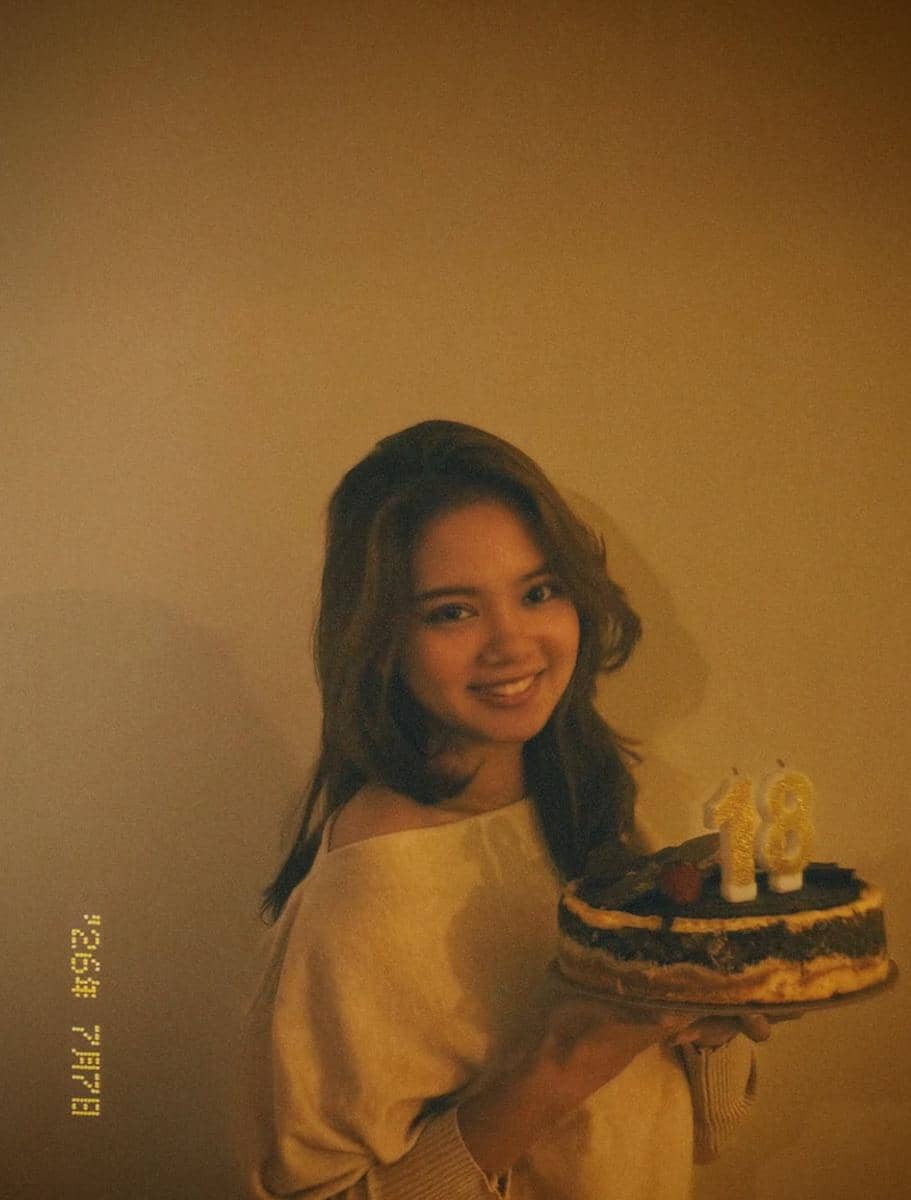 Seorang perempuan tersenyum sambil memegang kue ulang tahun dengan lilin angka 18 di atasnya dalam suasana hangat di dalam ruangan.