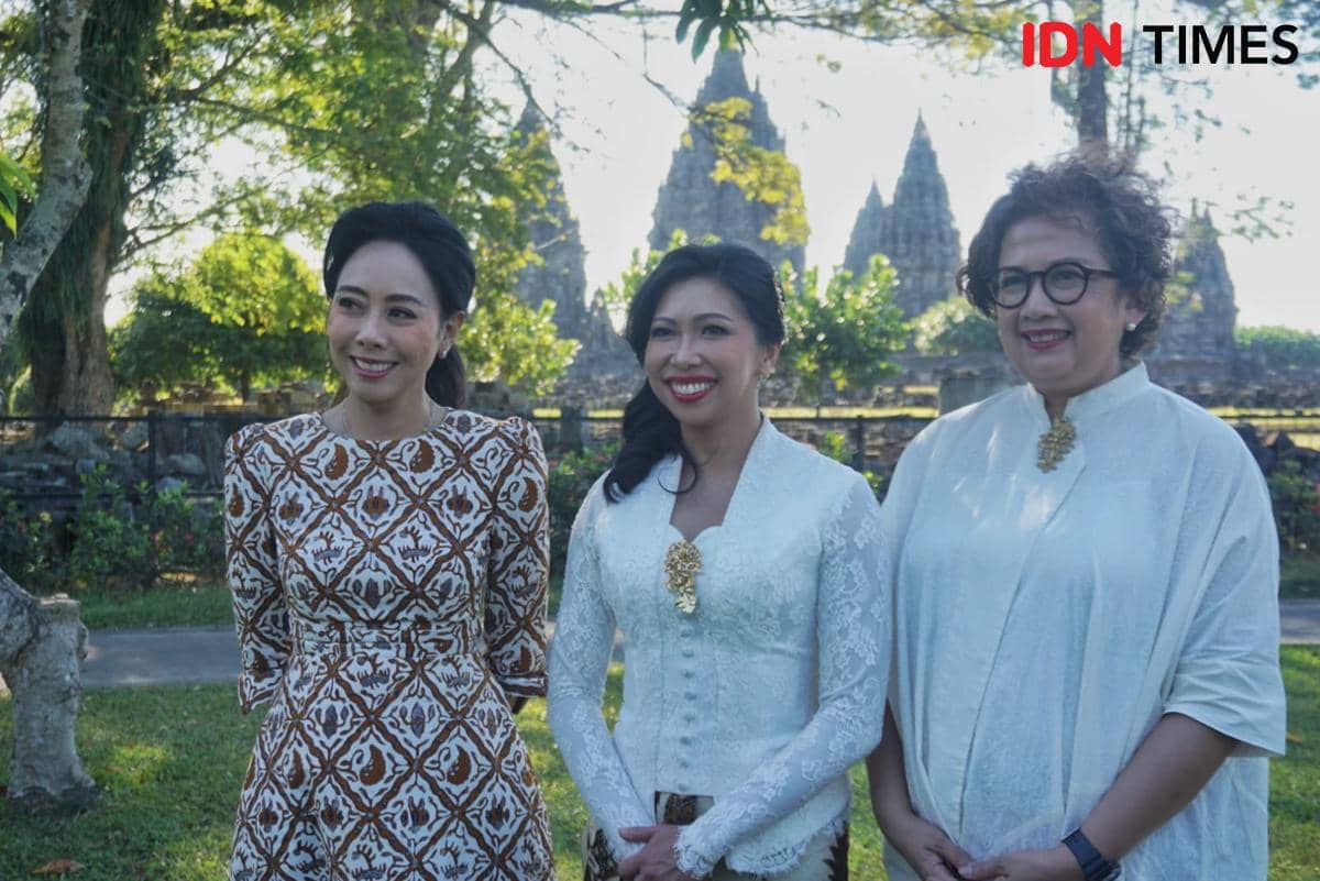 Tiga perempuan berbusana tradisional berdiri di area hijau dengan latar belakang Candi Prambanan yang tampak megah di kejauhan.
