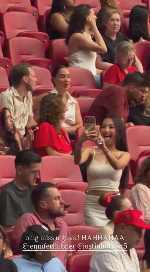 Seorang wanita mengenakan gaun putih tersenyum sambil mengambil foto di antara penonton konser yang duduk di kursi stadion berwarna merah.