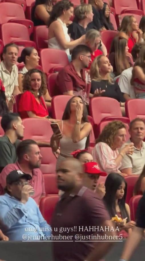 Seorang perempuan meniupkan ciuman sambil berdiri di antara penonton konser di stadion dengan kursi berwarna merah.