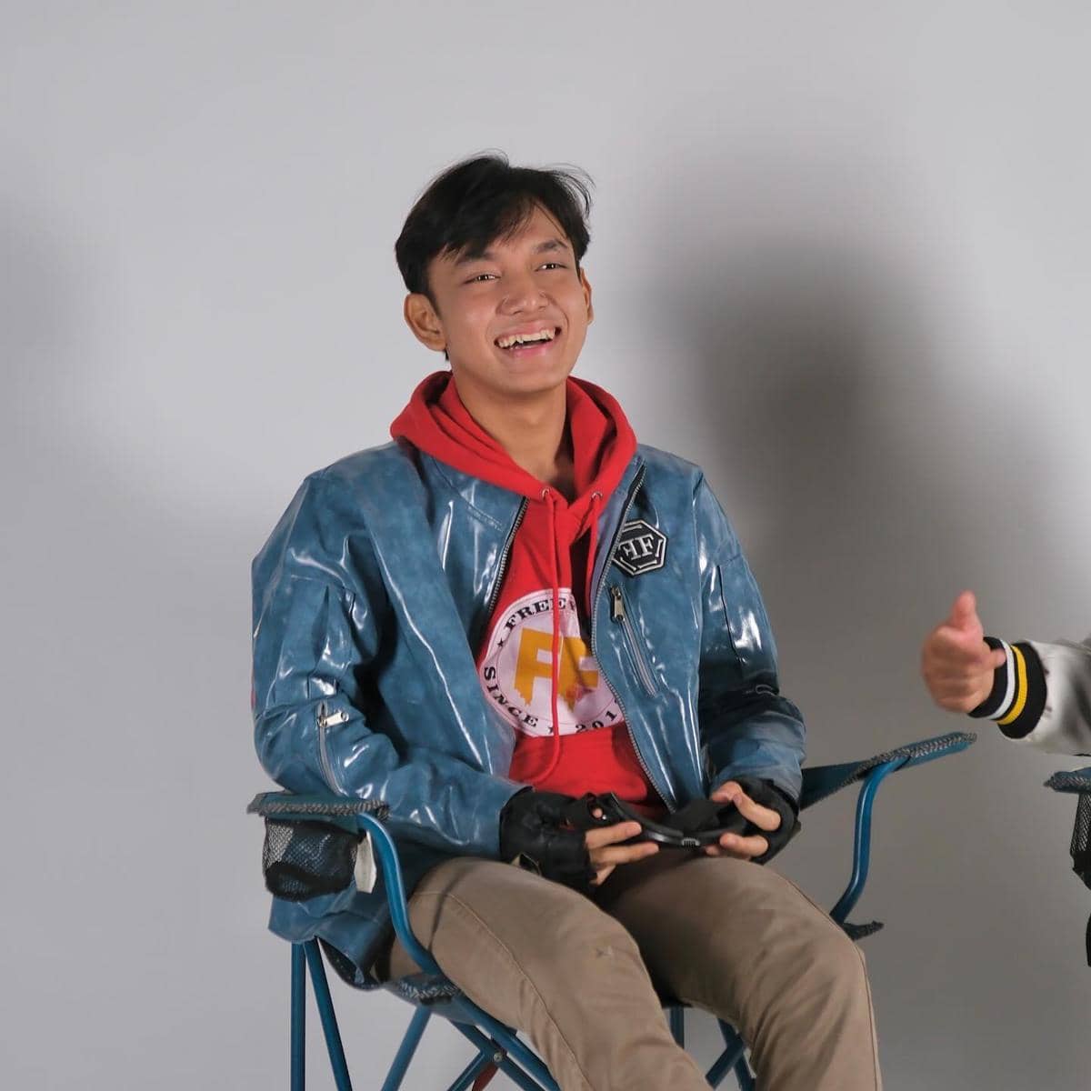 Seorang pria muda tersenyum sambil duduk di kursi lipat, mengenakan jaket biru mengilap dan hoodie merah dengan latar abu-abu polos.
