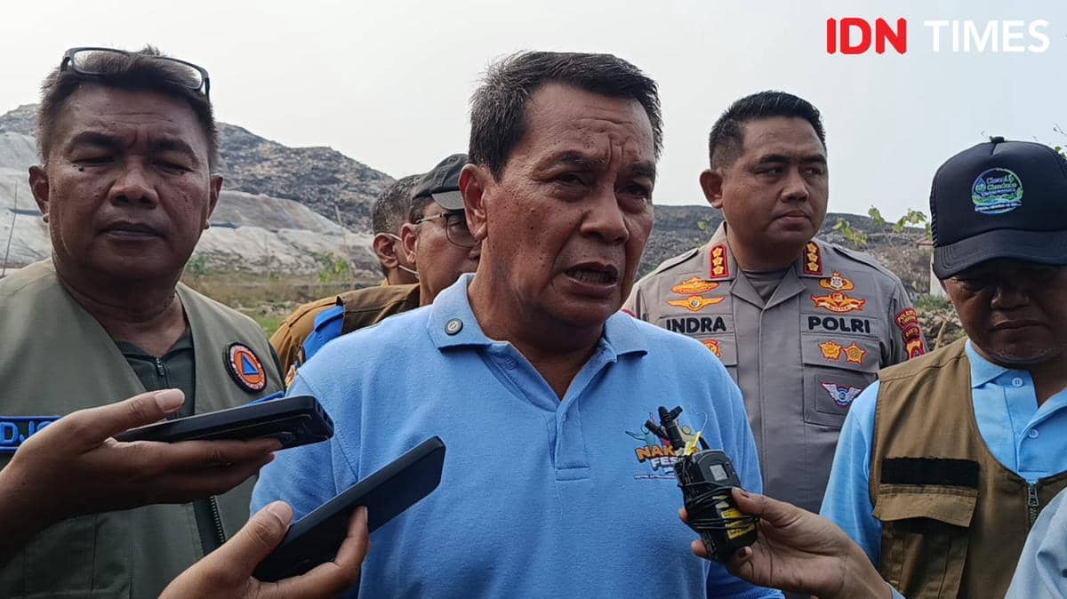 Seorang pria berbaju biru berbicara kepada wartawan sambil dikelilingi beberapa petugas berseragam di area perbukitan terbuka.