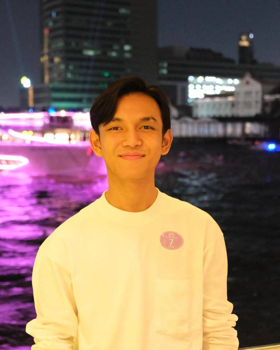Seorang pria muda mengenakan kaus putih tersenyum di malam hari dengan latar belakang kota dan cahaya ungu di tepi sungai.