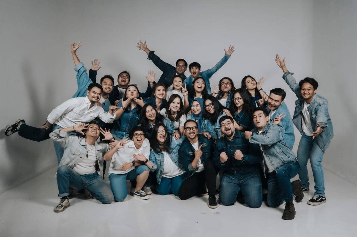 Sekelompok anggota tim IDN Event berpose ceria bersama di studio dengan latar putih, mengenakan pakaian bernuansa denim dan putih.