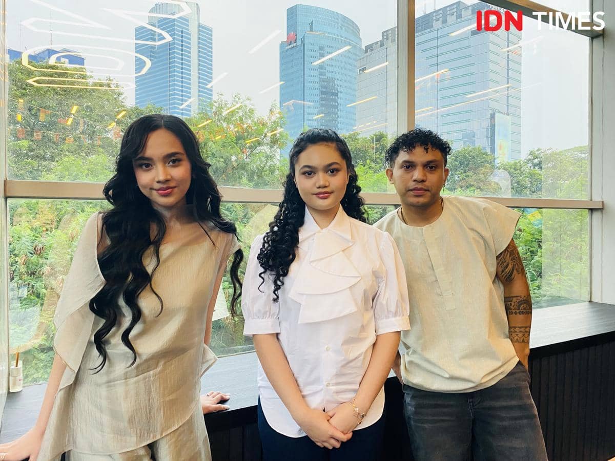 Tiga orang berdiri di depan jendela besar dengan latar gedung tinggi dan pepohonan di kantor IDN pada siang hari.