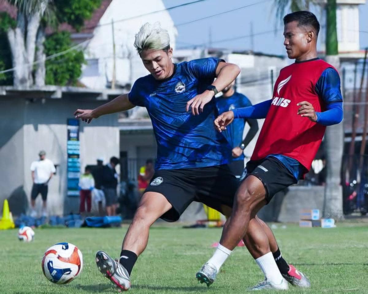 Dua pemain Arema FC berlatih di lapangan, salah satunya berusaha merebut bola saat sesi latihan tim di bawah sinar matahari.