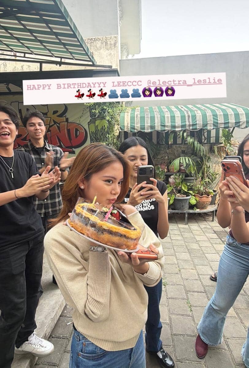 Seorang perempuan memegang kue ulang tahun dengan lilin menyala sambil berpose, dikelilingi teman-teman yang tersenyum dan bertepuk tangan.
