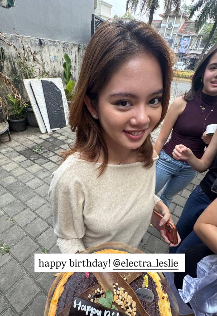 Seorang perempuan muda tersenyum sambil memegang kue ulang tahun di luar ruangan, dikelilingi oleh teman-temannya.