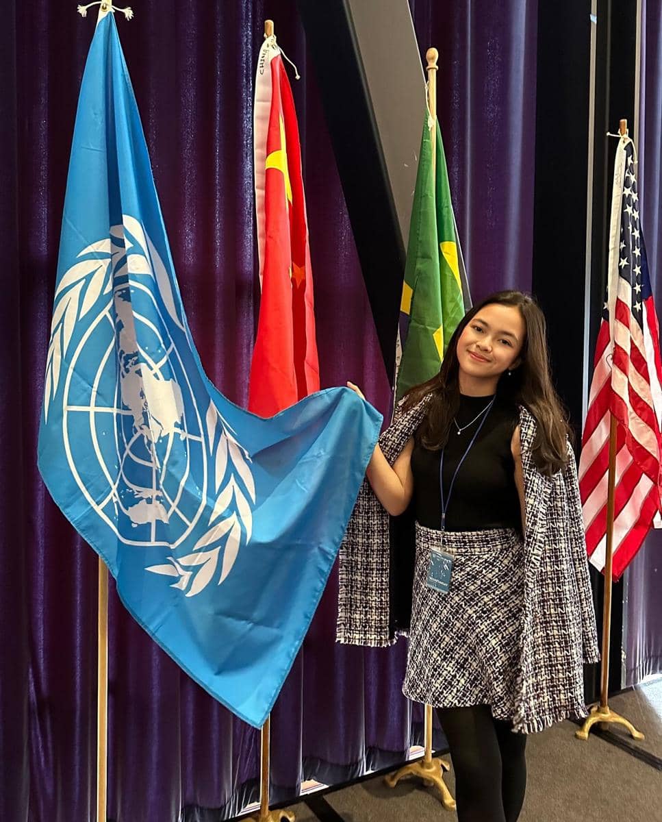 Seorang perempuan berdiri di samping bendera Perserikatan Bangsa-Bangsa dengan latar beberapa bendera negara di ruangan berwarna ungu.