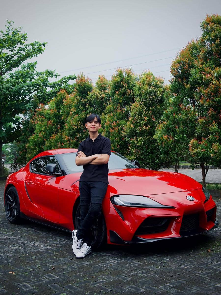 Seorang pria berdiri bersandar di mobil sport merah di area luar dengan latar pepohonan hijau dan jalan berbatu basah.
