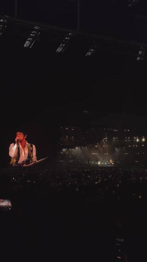 Panggung konser dengan penyanyi tampil di layar besar, dikelilingi cahaya dan penonton yang memenuhi arena pada malam hari.