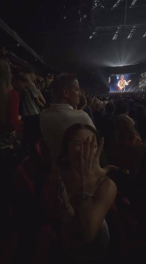 Seorang perempuan mengangkat tangan di tengah kerumunan penonton konser di stadion besar dengan panggung terang di kejauhan.