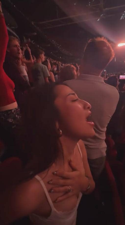 Seorang perempuan bernyanyi dengan penuh semangat di tengah kerumunan penonton konser di dalam arena yang dipenuhi cahaya panggung.