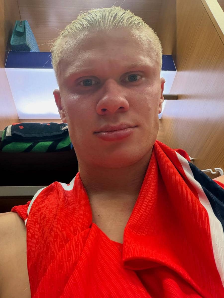 Seorang pria berambut pirang mengenakan jersey merah berpose selfie di dalam ruang ganti dengan latar kayu dan handuk di belakangnya.