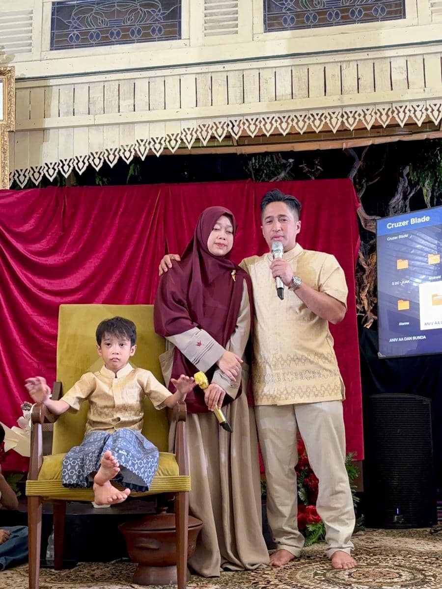 Seorang pria dan wanita berdiri berdampingan mengenakan busana tradisional dengan seorang anak kecil duduk di kursi di depan tirai merah.