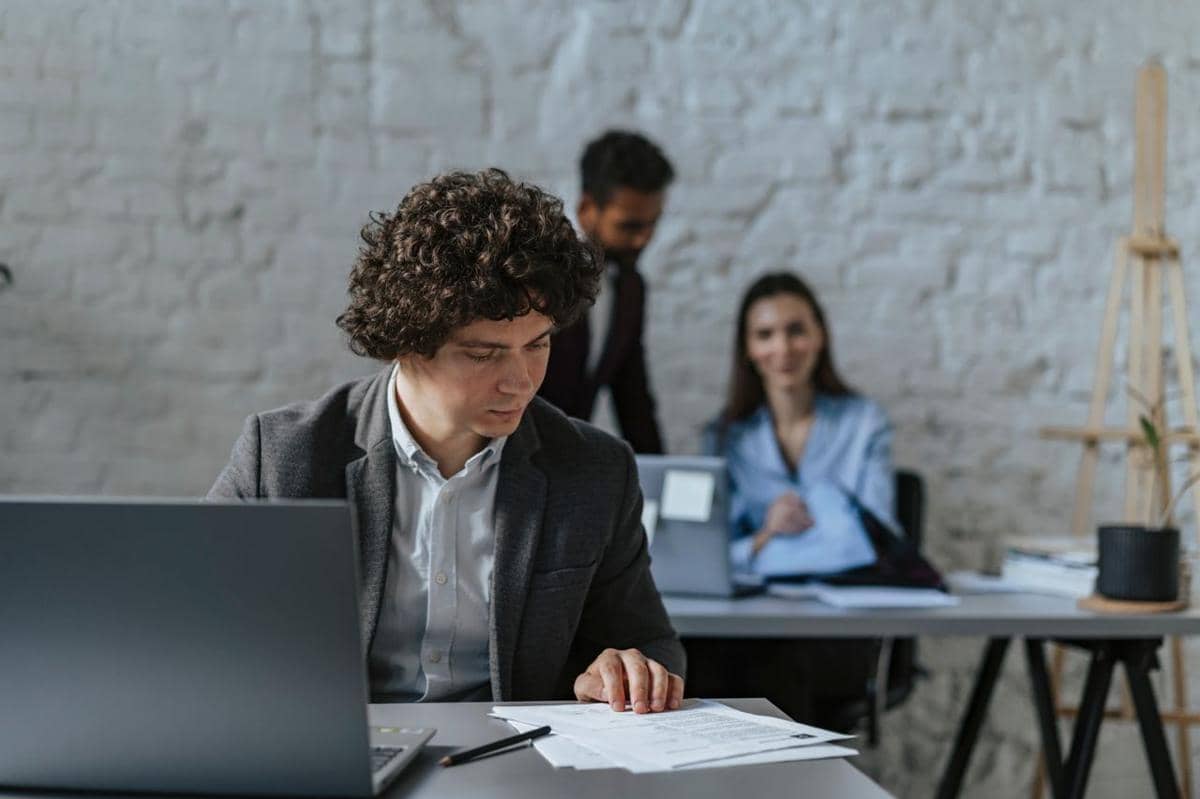 Seorang pria mengenakan jas bekerja di meja dengan laptop dan dokumen, sementara dua rekan kerja berbincang di latar belakang kantor.