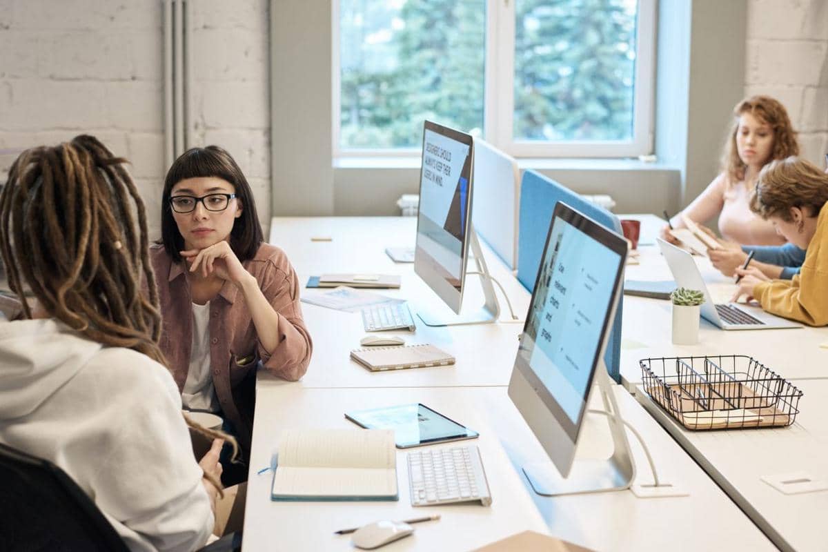Beberapa orang bekerja dan berdiskusi di ruang kantor modern dengan komputer, buku catatan, dan suasana kolaboratif.