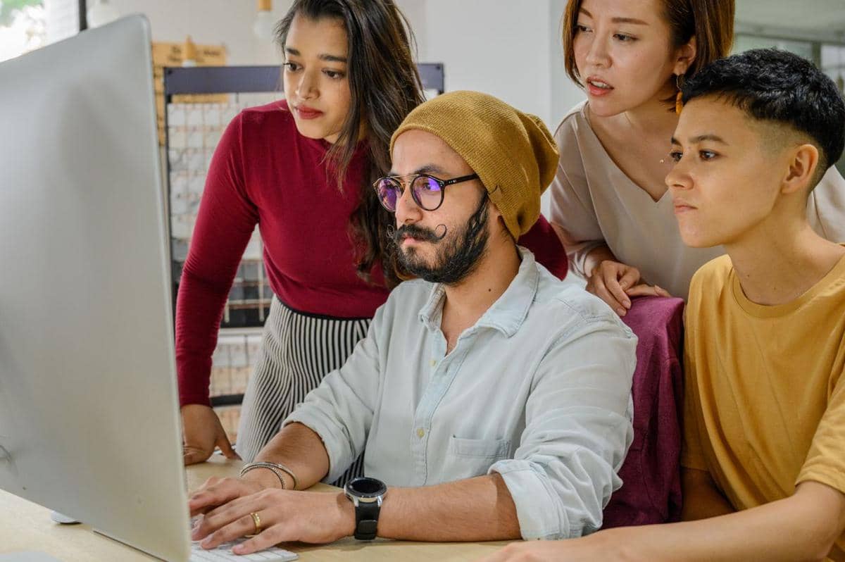 Empat orang bekerja sama di depan komputer di kantor, memperhatikan layar dengan serius sambil berdiskusi tentang pekerjaan.
