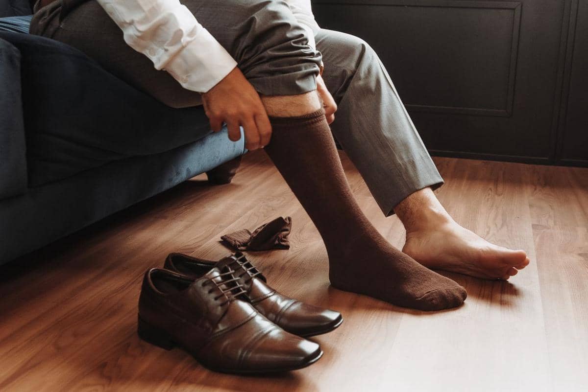 Seorang pria duduk di sofa sedang mengenakan kaus kaki cokelat dan bersiap memakai sepatu kulit cokelat di lantai kayu.