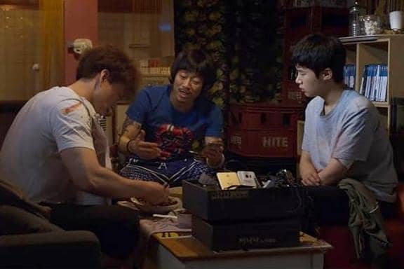 Tiga remaja duduk di ruang kecil dengan pencahayaan redup, tampak berdiskusi serius sambil memegang uang dan peralatan elektronik.
