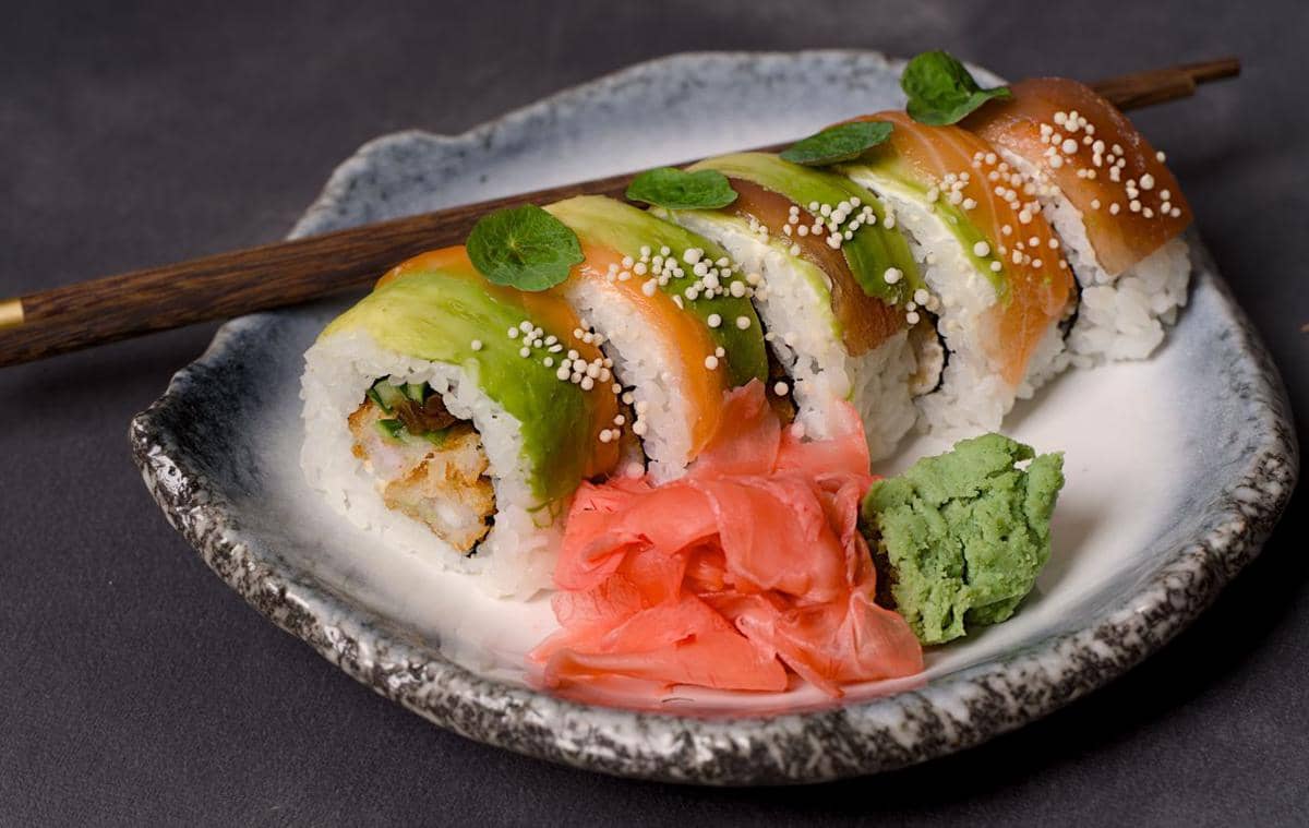 Beberapa potong sushi gulung dengan topping alpukat dan ikan disajikan di piring keramik bersama wasabi dan jahe merah acar.
