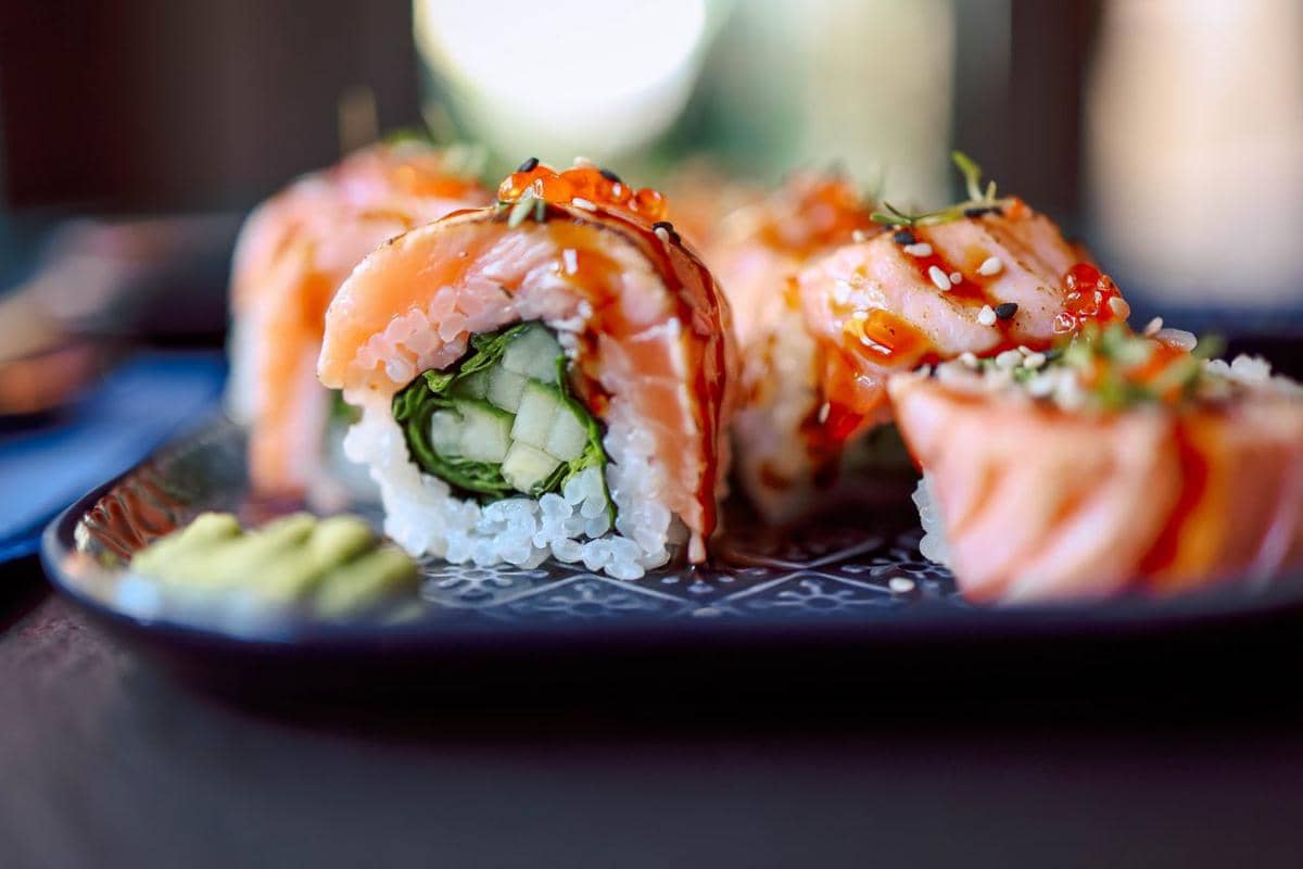 Beberapa potong sushi dengan isian sayuran dan ikan salmon segar disajikan di atas piring hitam dengan saus dan taburan wijen.