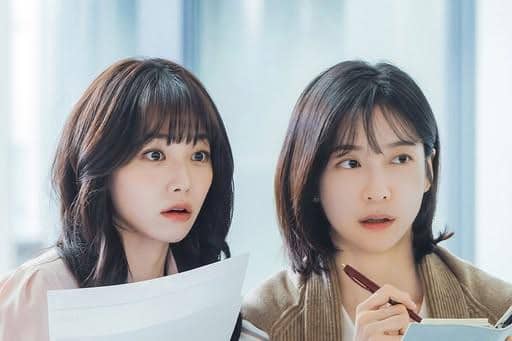 Dua wanita muda memegang dokumen dan pena sambil menatap ke arah yang sama dengan ekspresi terkejut di kantor modern.