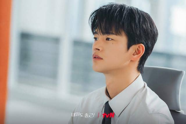 Seorang pria muda mengenakan kemeja putih dan dasi hitam duduk di kursi kantor dengan ekspresi serius di ruang kerja modern.
