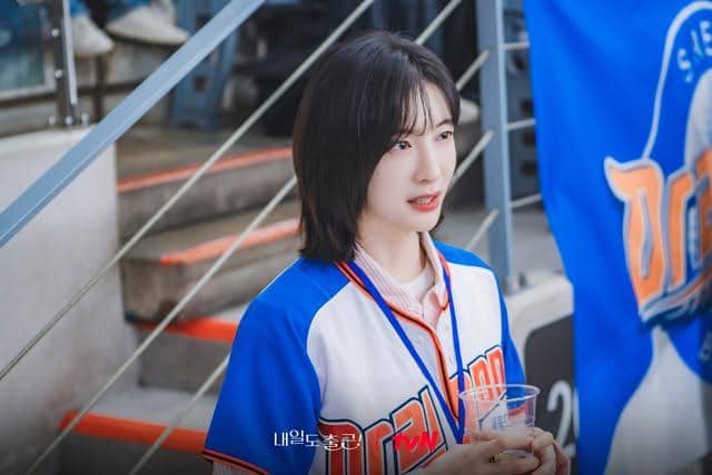 Seorang wanita mengenakan seragam bisbol biru putih sedang berdiri di tribun stadion sambil memegang gelas plastik.