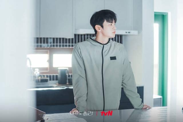 Seorang pria mengenakan jaket abu-abu berdiri di dapur modern dengan pencahayaan lembut, tampak menatap ke arah jendela.