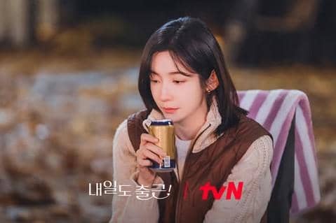 Seorang wanita berambut pendek mengenakan jaket cokelat duduk sambil memegang minuman kaleng di lokasi syuting drama See You at Work Tomorrow.
