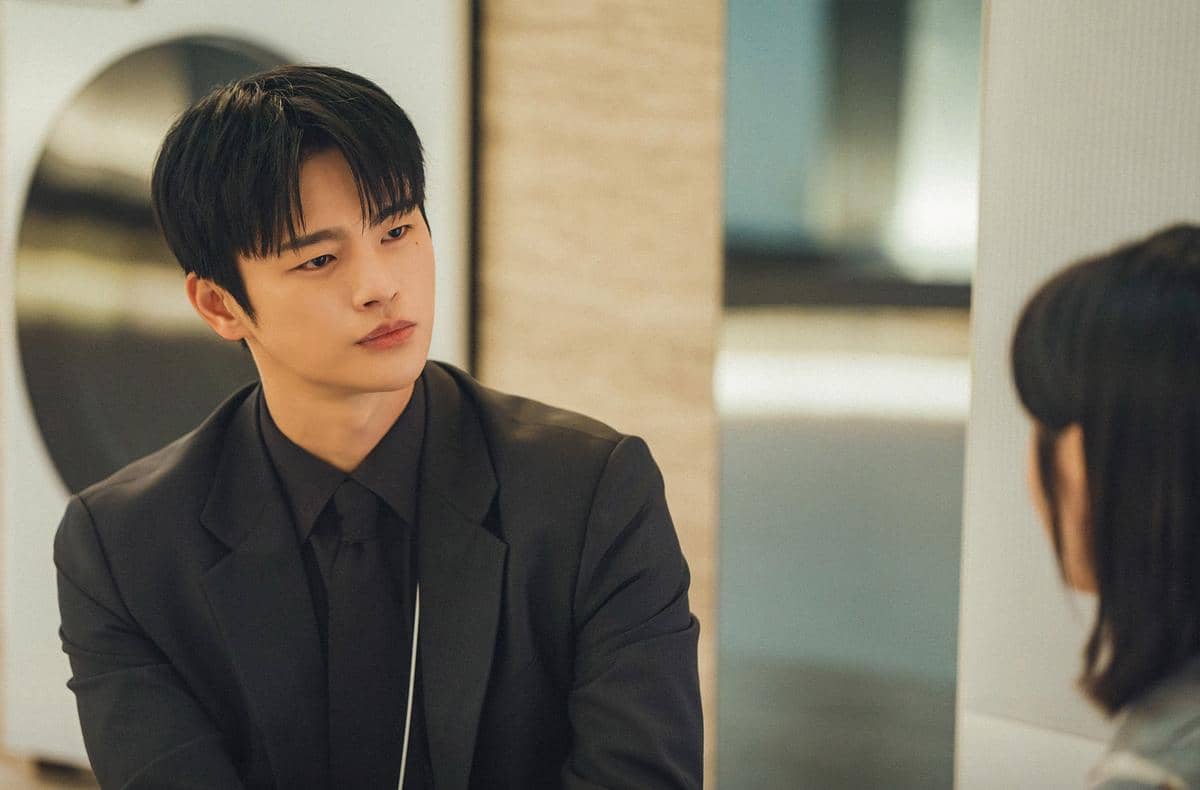 Seorang pria mengenakan setelan jas hitam duduk dan menatap seseorang di depannya dalam adegan drama Korea See You at Work Tomorrow.