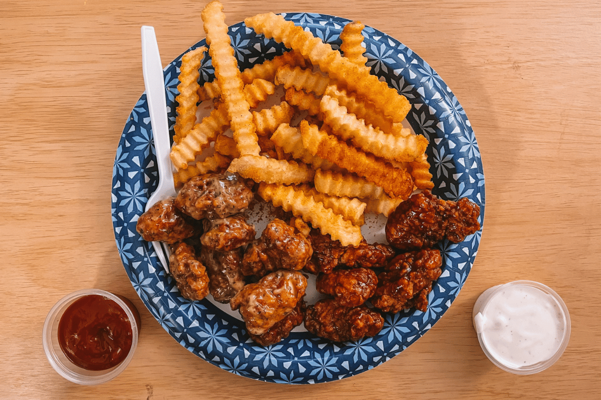 Sepiring dakgangjeong kaki lima disajikan dengan kentang goreng bergelombang, saus merah, dan saus putih di atas meja kayu.