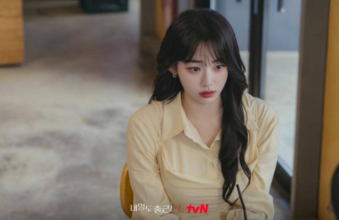Seorang perempuan berambut panjang mengenakan kemeja kuning duduk di dalam ruangan dengan ekspresi serius dalam cuplikan drama See You at Work Tomorrow.