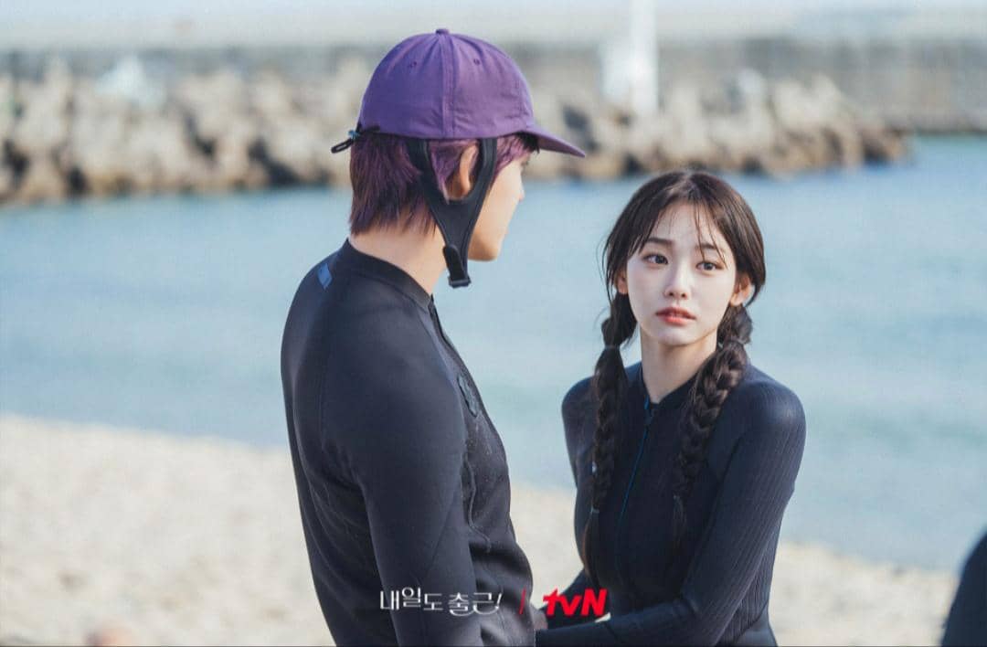 Dua orang mengenakan pakaian selam hitam berbicara di tepi pantai dengan latar laut dan pemecah ombak dalam cuplikan drama See You at Work Tomorrow.
