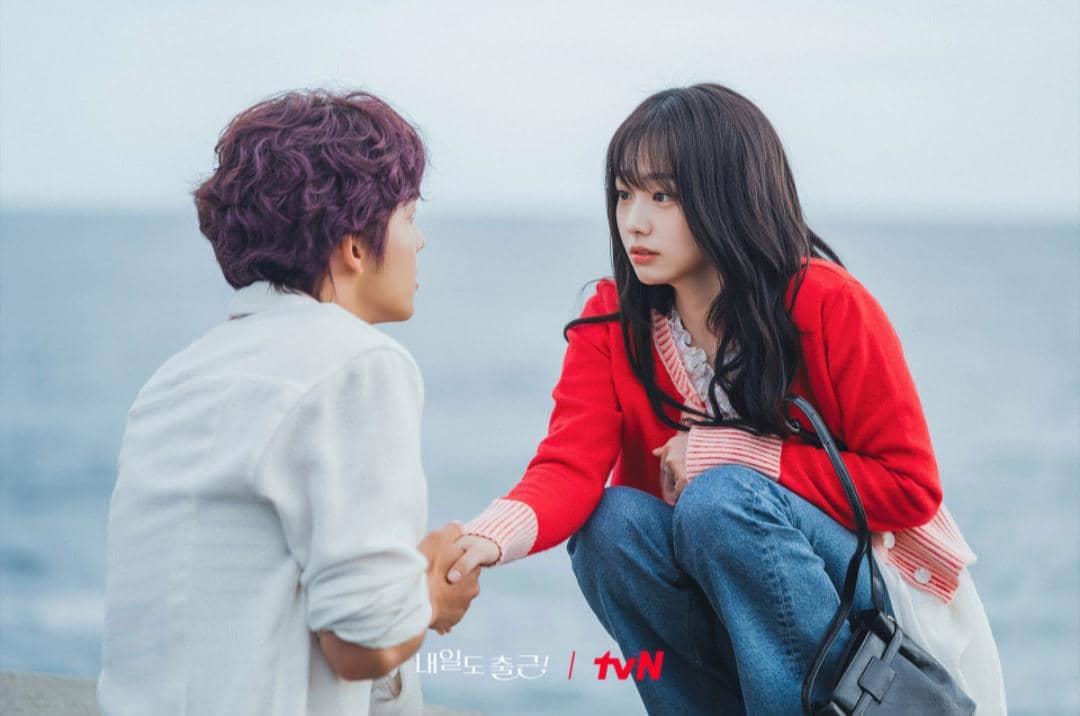 Dua karakter dalam drama See You at Work Tomorrow duduk di tepi laut, saling menatap dan berpegangan tangan dengan ekspresi serius.
