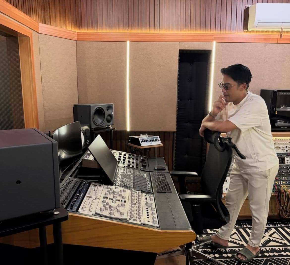 Seorang pria mengenakan pakaian kasual berdiri di ruang studio musik modern, memperhatikan peralatan audio dan layar komputer di depannya.