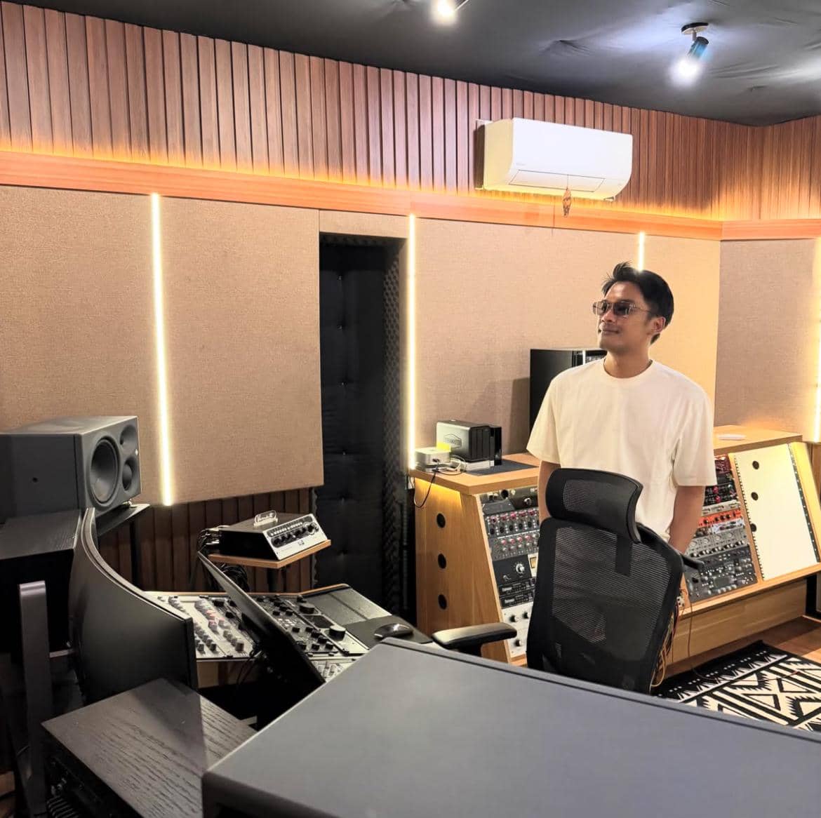 Seorang pria mengenakan kaus putih berdiri di dalam studio rekaman modern dengan peralatan audio profesional di sekelilingnya.