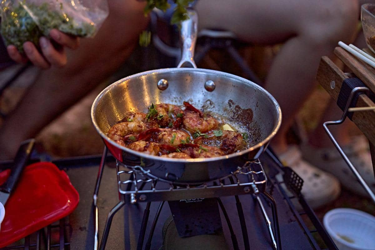 Wajan berisi ayam goreng saus manis pedas sedang dimasak di atas kompor portabel dengan taburan daun hijau segar.