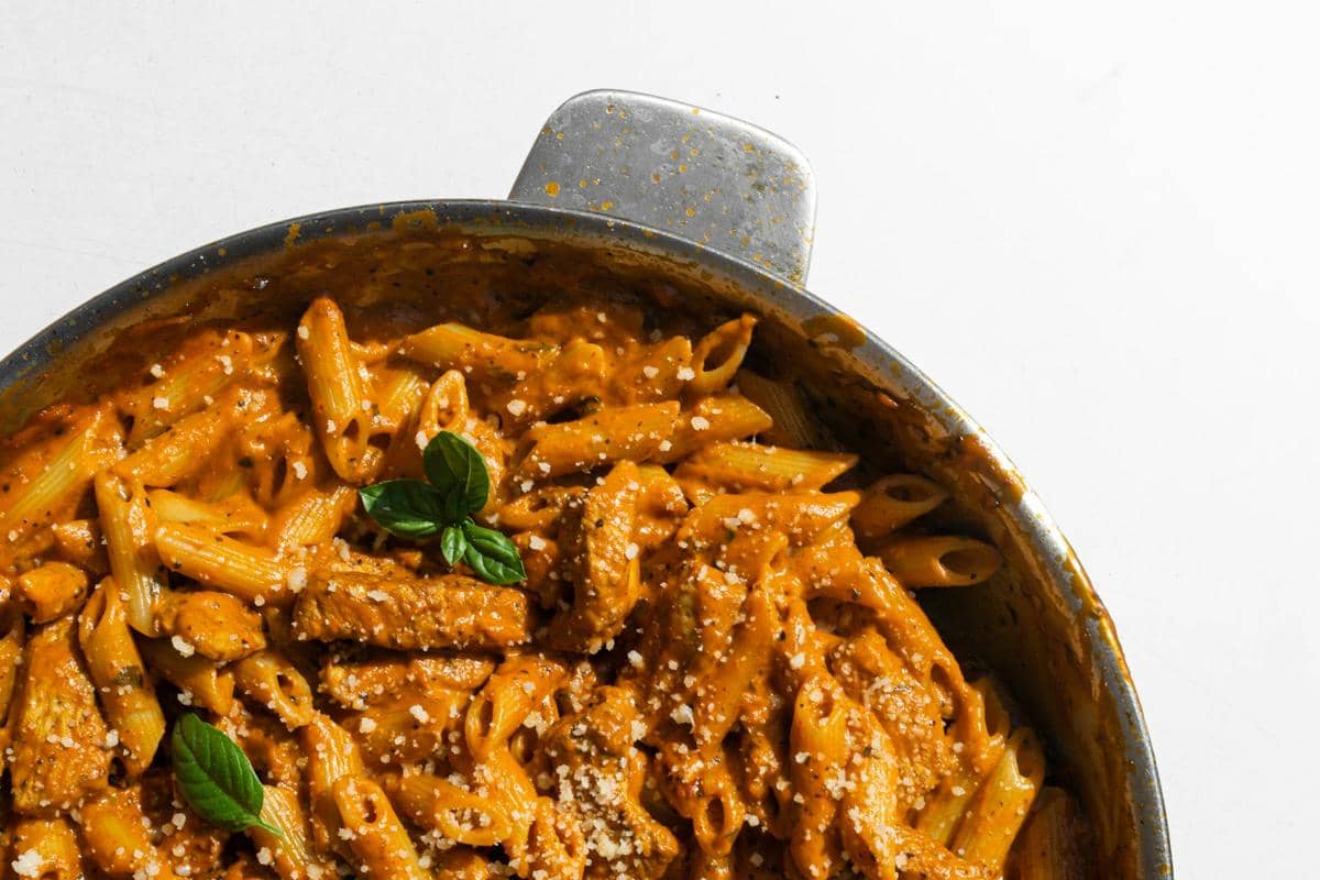 Pasta penne dengan saus gochujang alfredo berwarna oranye krem disajikan dalam wajan logam, dihiasi daun basil segar dan taburan keju parmesan.