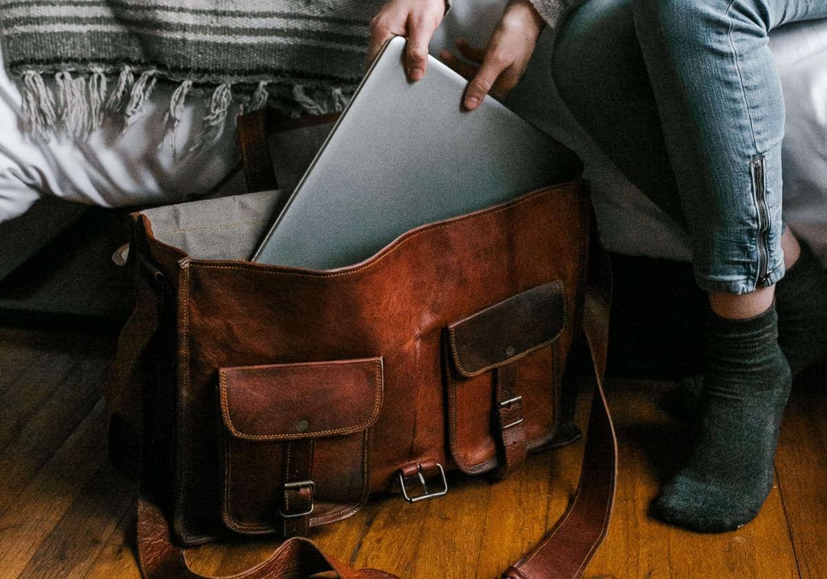 Seseorang memasukkan laptop ke dalam tas kerja kulit berwarna cokelat di atas lantai kayu dekat tempat tidur.