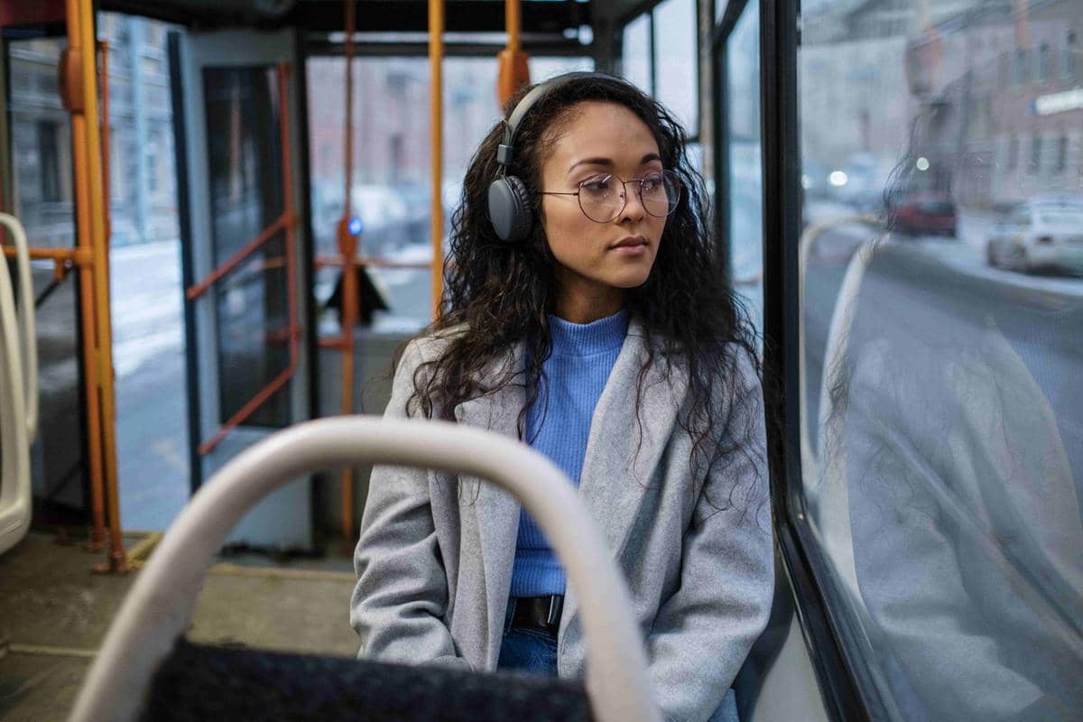 Seorang perempuan mengenakan headphone dan kacamata duduk di dalam bus sambil melihat ke luar jendela di siang hari.