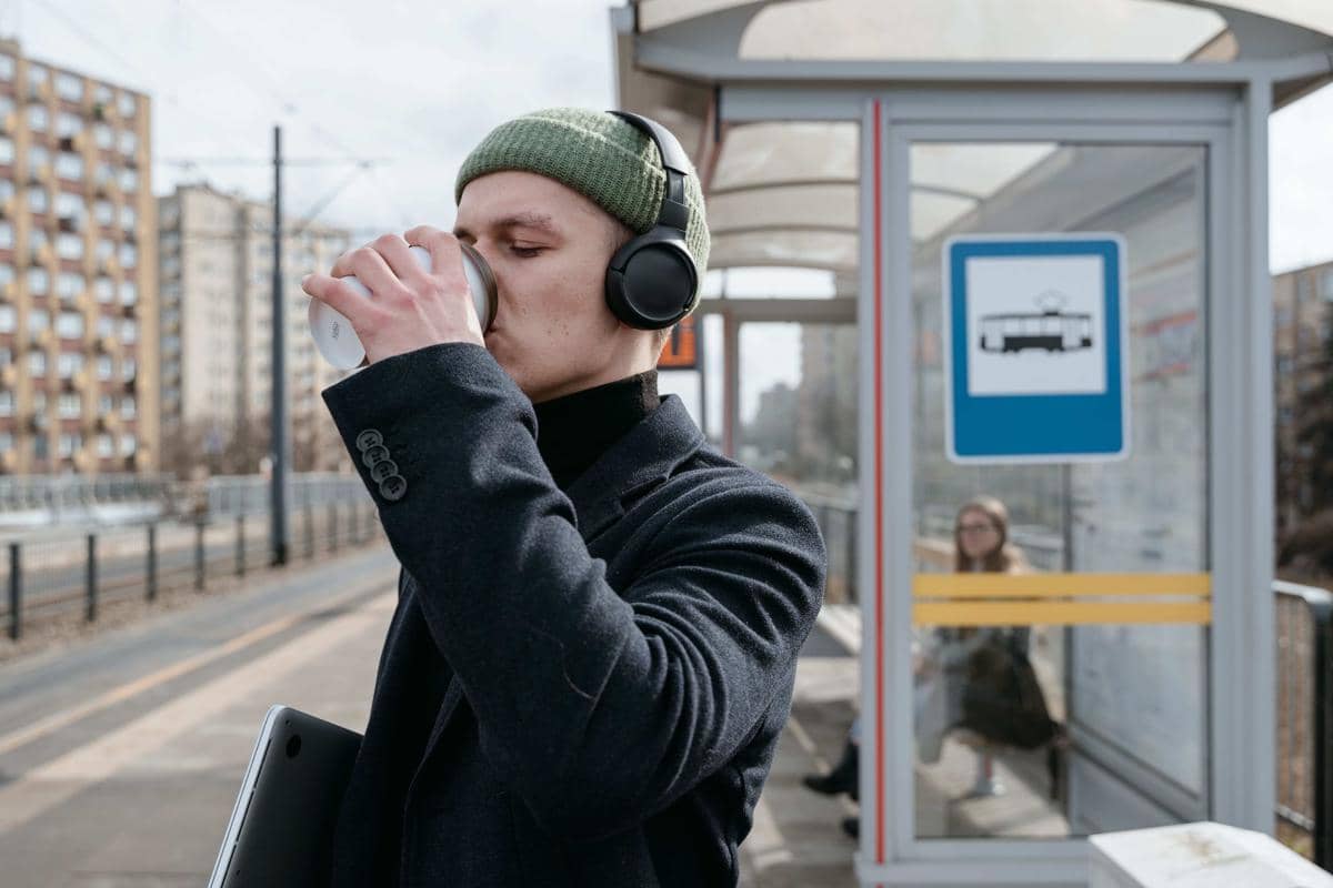 Seorang pria mengenakan beanie hijau dan headphone hitam sedang minum dari gelas logam di halte transportasi umum.