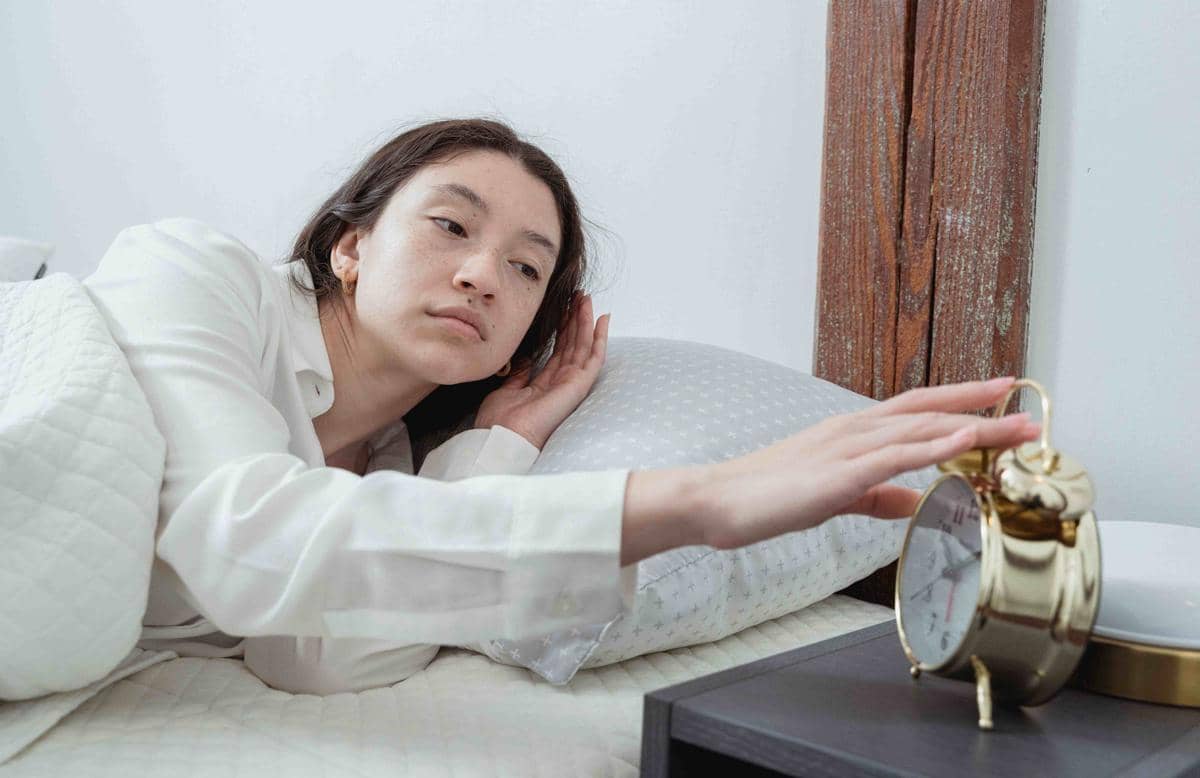 Seorang wanita tampak mengantuk berbaring di tempat tidur sambil meraih jam alarm berwarna emas di meja samping tempat tidurnya.