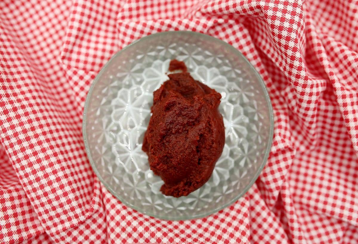 Semangkuk bumbu merah kental di atas kain bermotif kotak merah putih, disajikan dalam mangkuk kaca berpola.